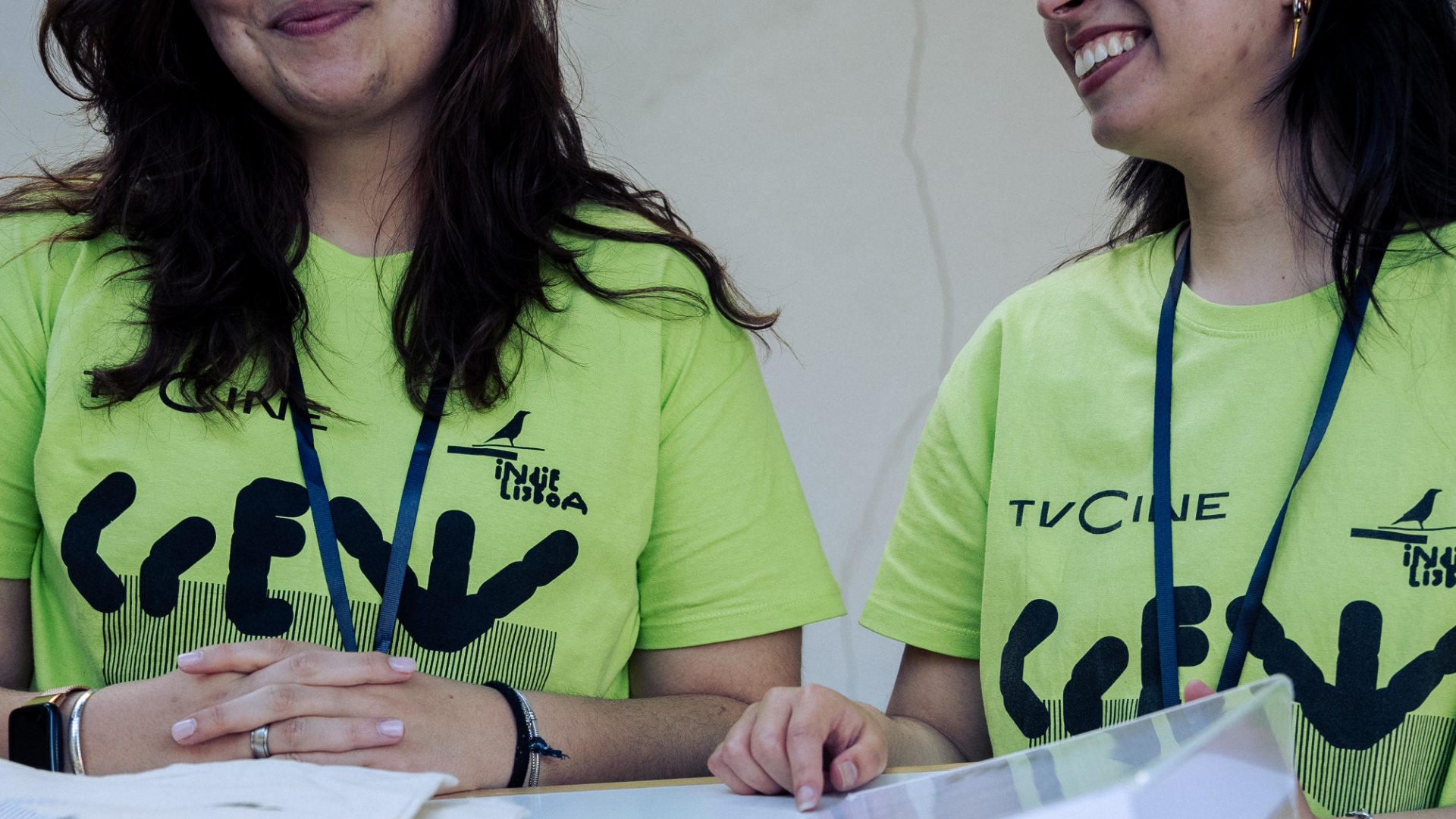 Duas voluntárias com t-shirts verdes do festival atendem participantes numa mesa de apoio