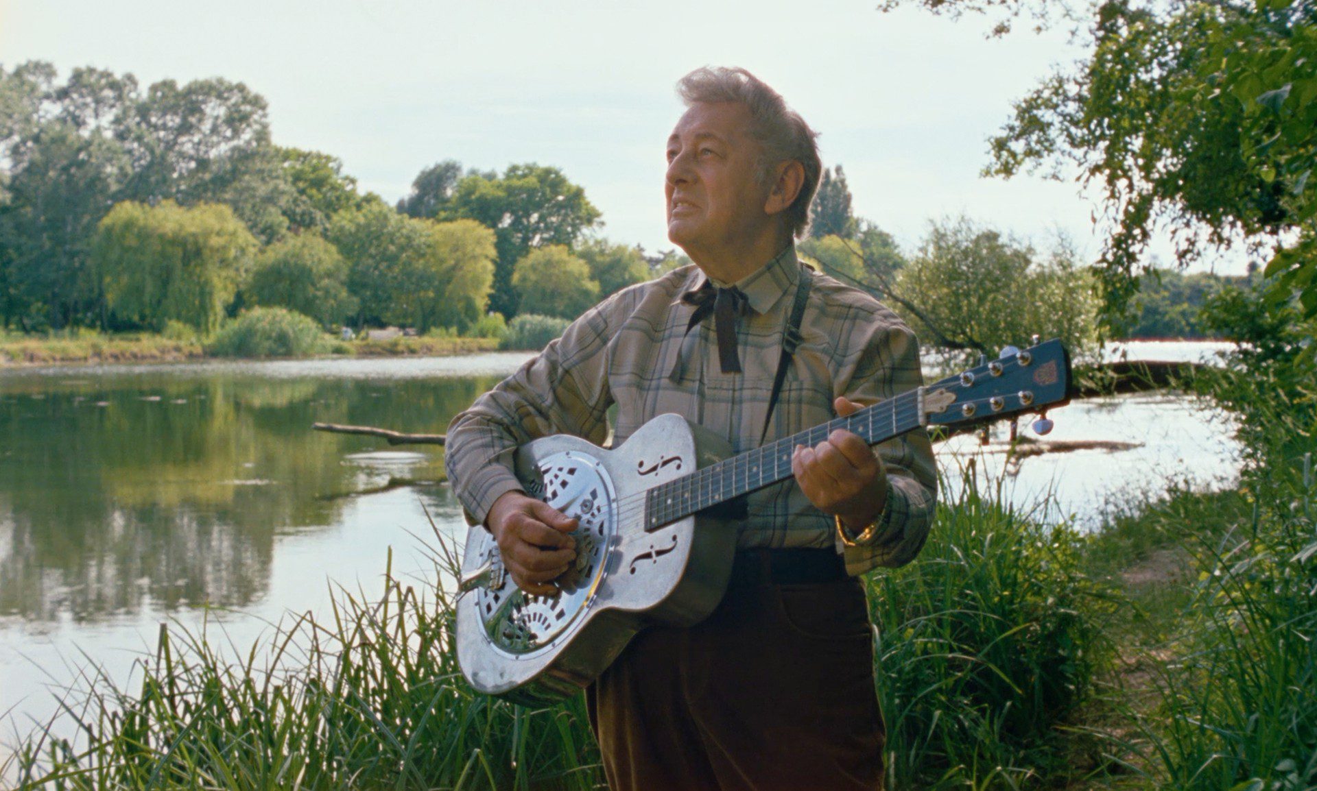 The Opening and Closing Ceremonies of IndieLisboa 2026 1 A man plays the guitar by a lake, surrounded by greenery, looking up as he sings outdoors in a peaceful setting.