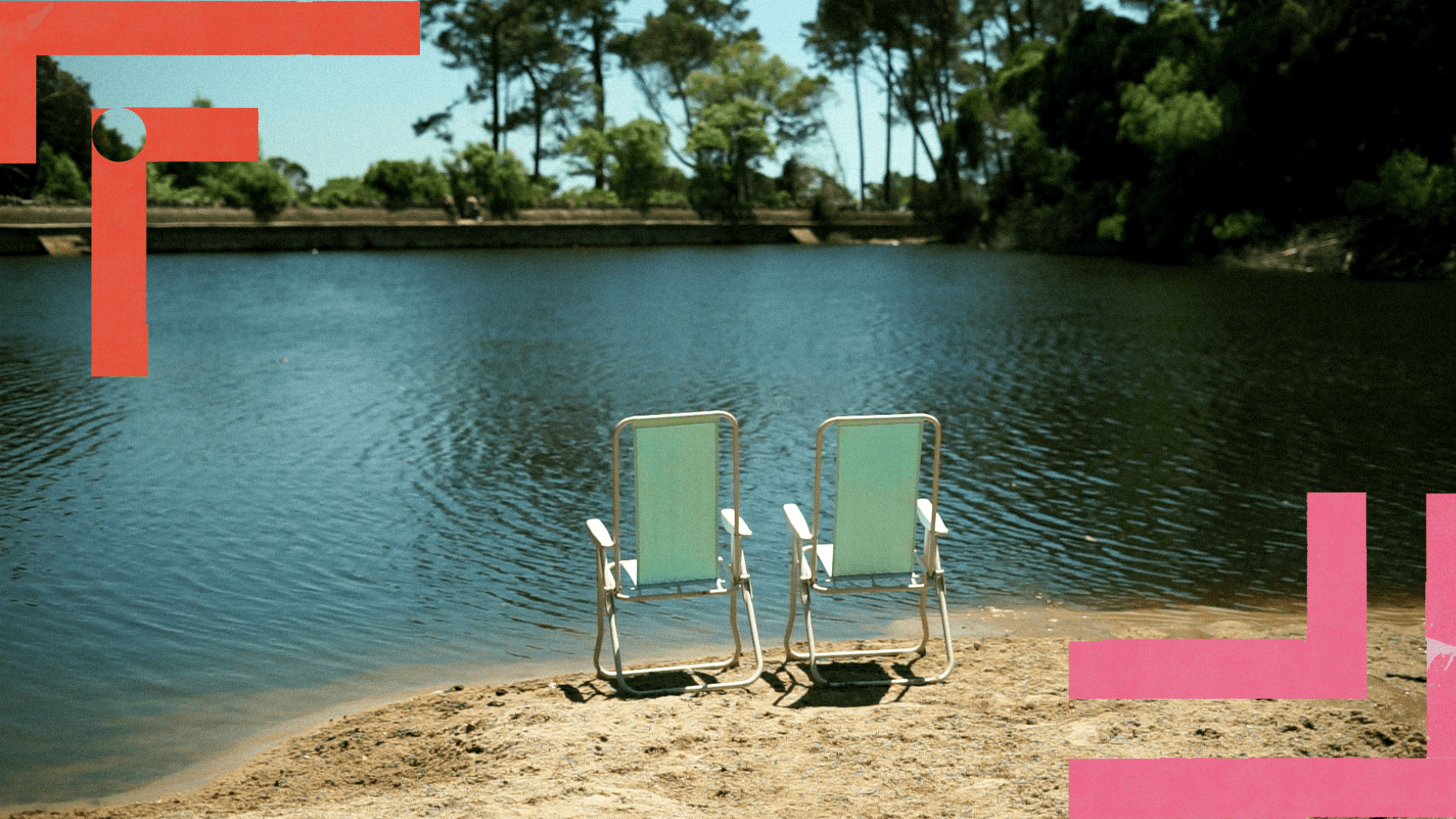 Duas cadeiras de praia vazias, lado a lado, estão posicionadas na margem arenosa de um lago calmo, voltadas para a água. A superfície do lago reflete a luz do dia e é cercada por árvores altas e vegetação densa ao fundo. A cena transmite tranquilidade e contemplação.